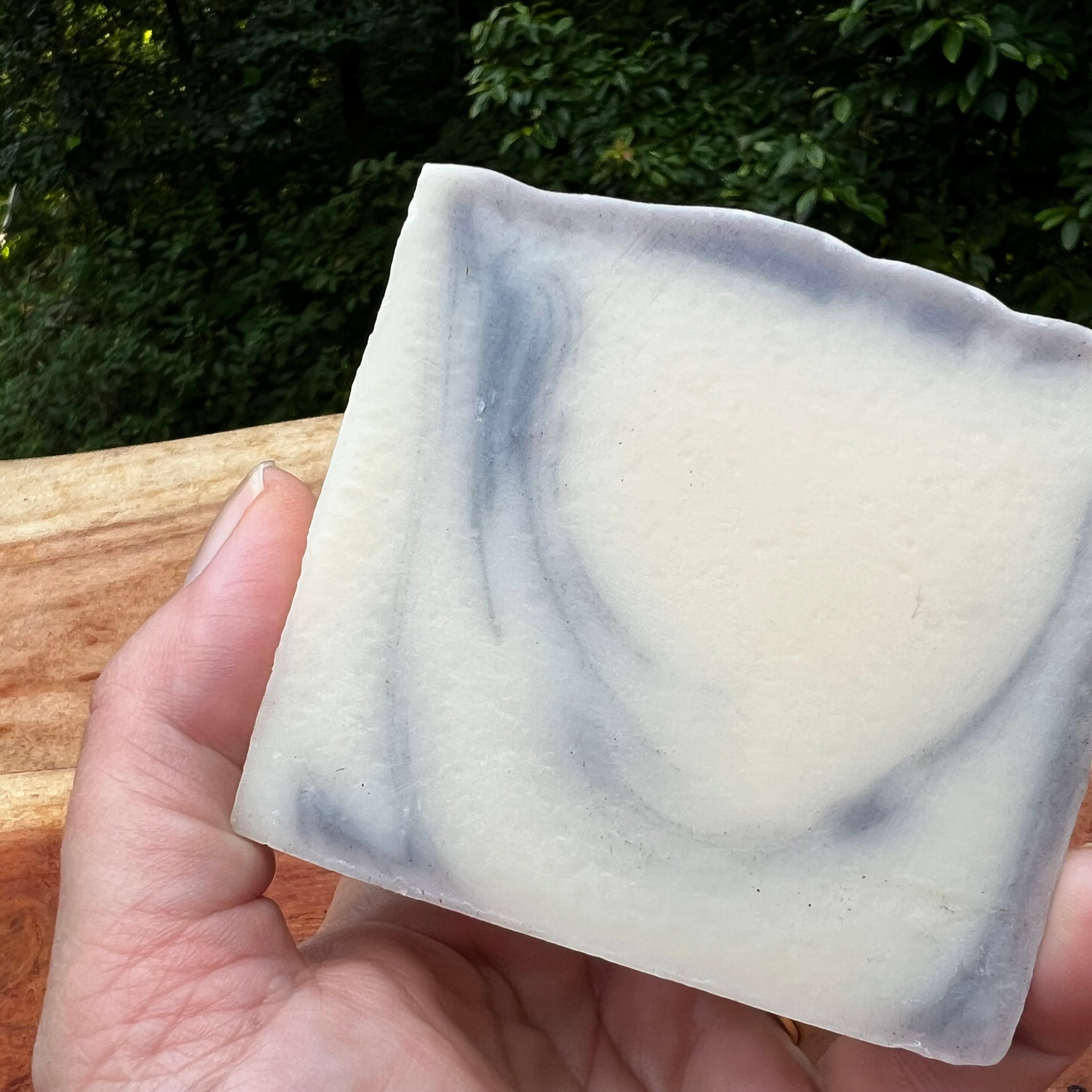 A hand holding a rectangular bar of handmade soap with a marbled cream and slate-gray pattern, set against a background of green foliage and a wooden railing.