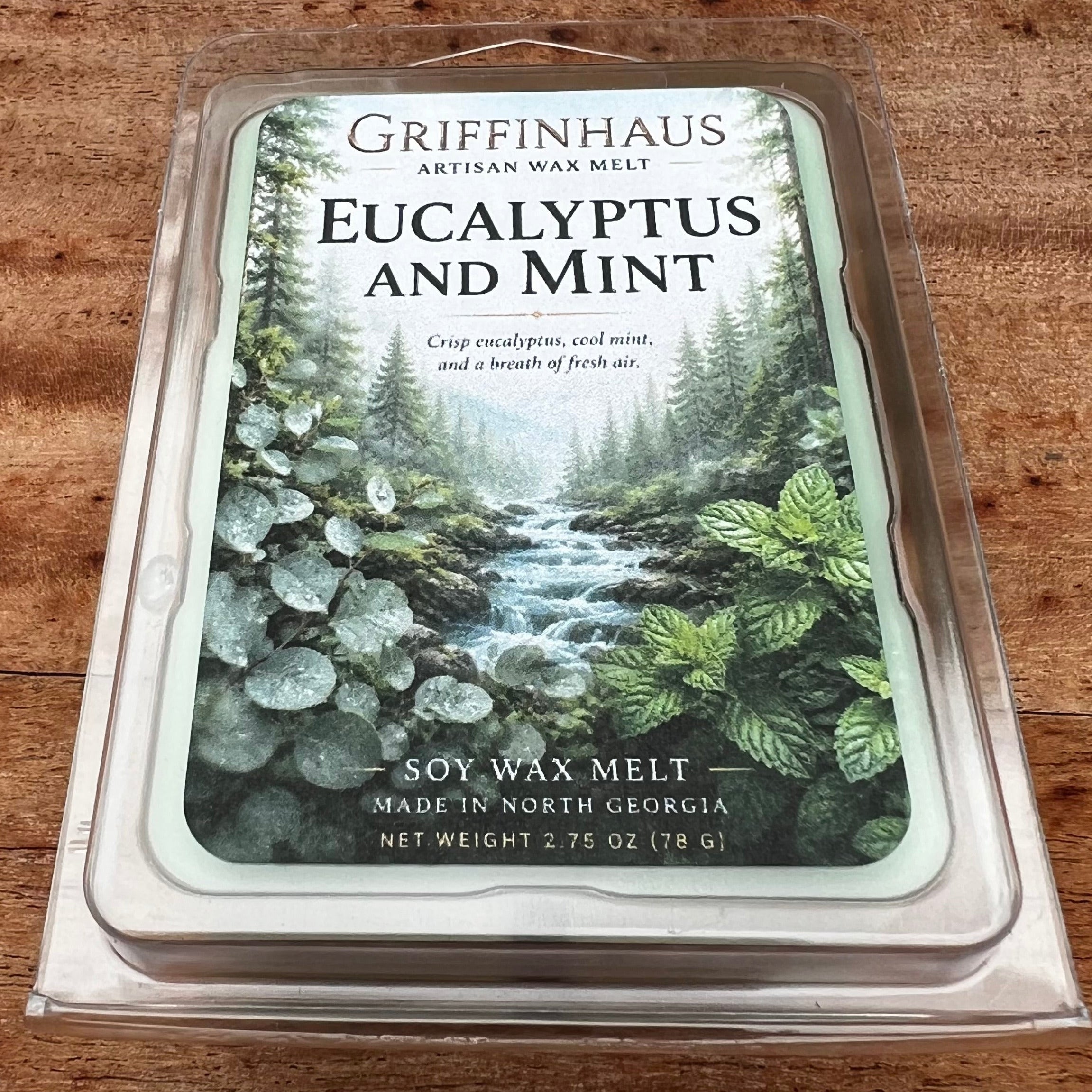 Griffinhaus Eucalyptus and Mint soy wax melt in a clear plastic clamshell package, displayed on a rustic wooden surface. The label features a forest illustration with a stream and green foliage.