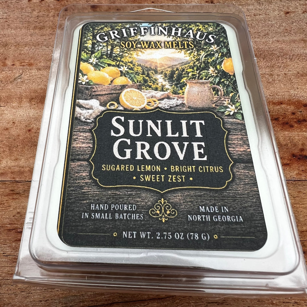 Griffinhaus Sunlit Grove soy wax melts in a clear plastic clamshell package, placed on a wooden surface. The label features a dark wood-grain background with illustrations of lemons, a mug, and a scenic landscape.